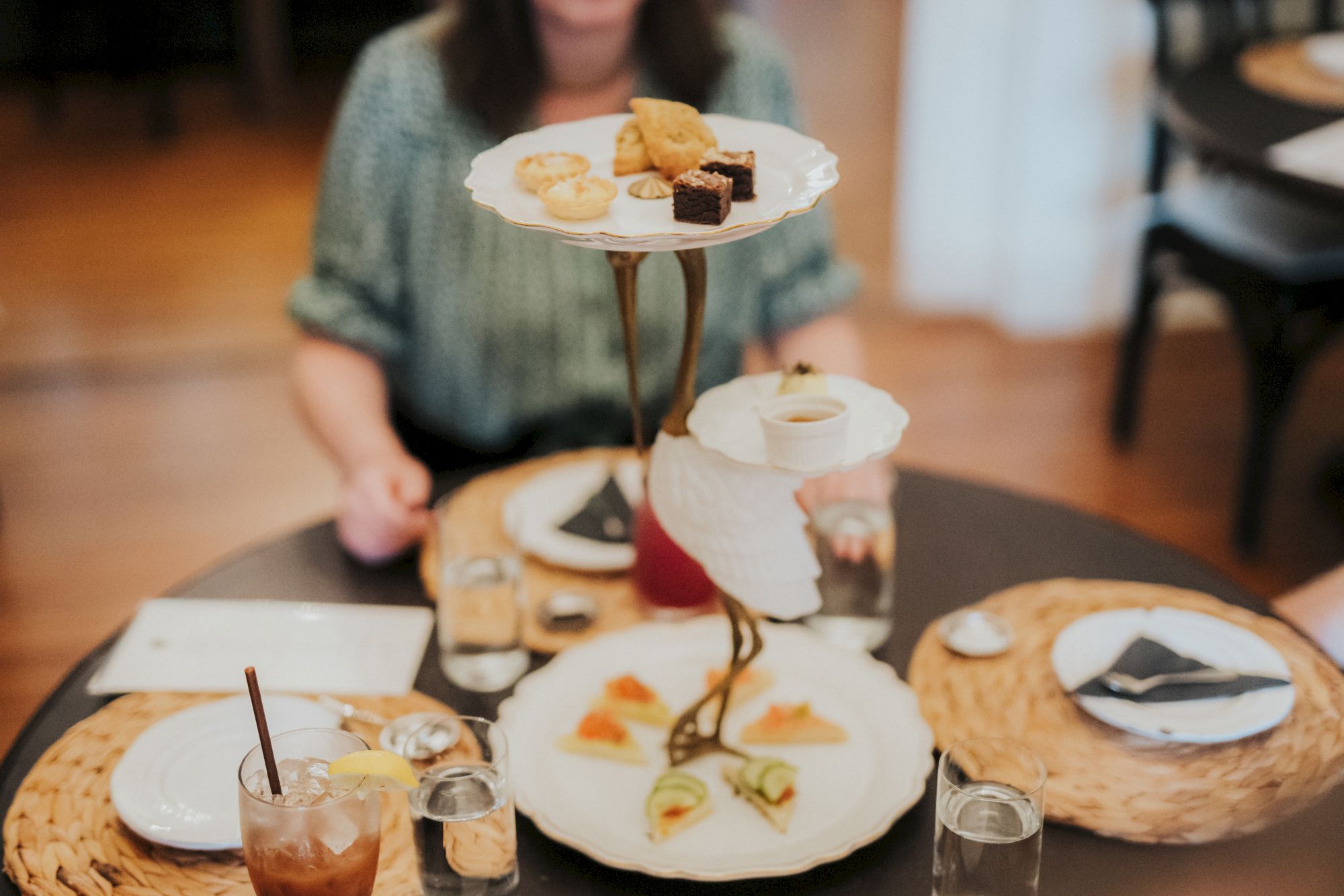 A table set for high tea with tiered trays of pastries and sandwiches, two glasses, and beverages, with a person blurred in the background.